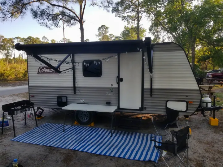 This inviting RV rental campsite features a grey travel trailer with a lit awning, outdoor cooking setup, chairs, and a striped mat on the dirt ground amidst trees.. Coleman Lantern 17B 2023