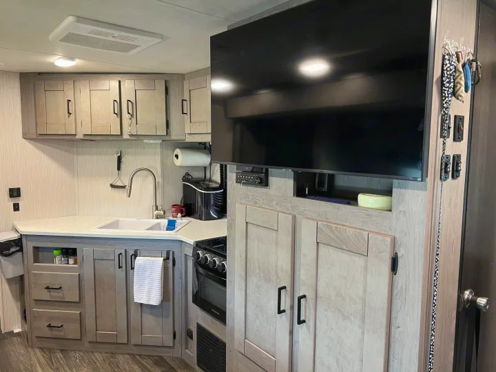 Inside an RV rental: a compact kitchen with light wood cabinets, sink, stove, TV, and pet leashes hanging on the wall.. Heartland RVs Mallard 2021