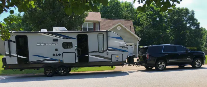 Black SUV hitched to a large grey travel trailer (Cherokee Alpha Wolf) parked on a residential street. Consider an rv rental!. Forest River Cherokee Alpha Wolf 2022