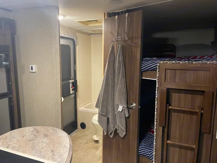 Inside an RV rental, showing a compact bathroom, towels hanging, and bunk beds with striped bedding.. Coachmen Apex 2017