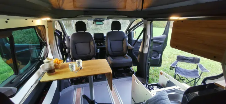 Cozy RV rental interior featuring a wooden dining table, kitchen area, front seating, and scenic outdoor views.. Renault Trafic 2023