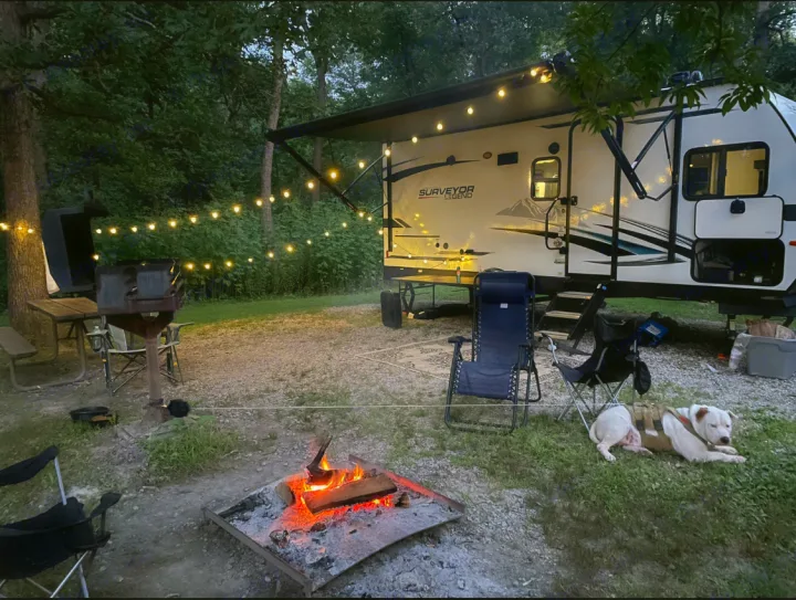 Cozy RV rental campsite with a warm campfire, string lights, and a relaxed dog. Perfect for an evening under the stars.. Forest River Surveyor Legend 2023