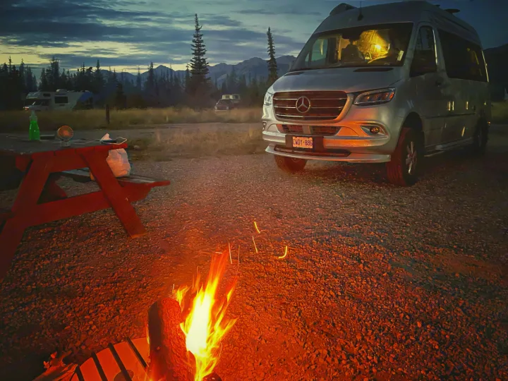 An RV rental campervan parked at a mountain campsite, illuminated by a glowing campfire under a beautiful dusk sky.. Airstream Interstate 2022