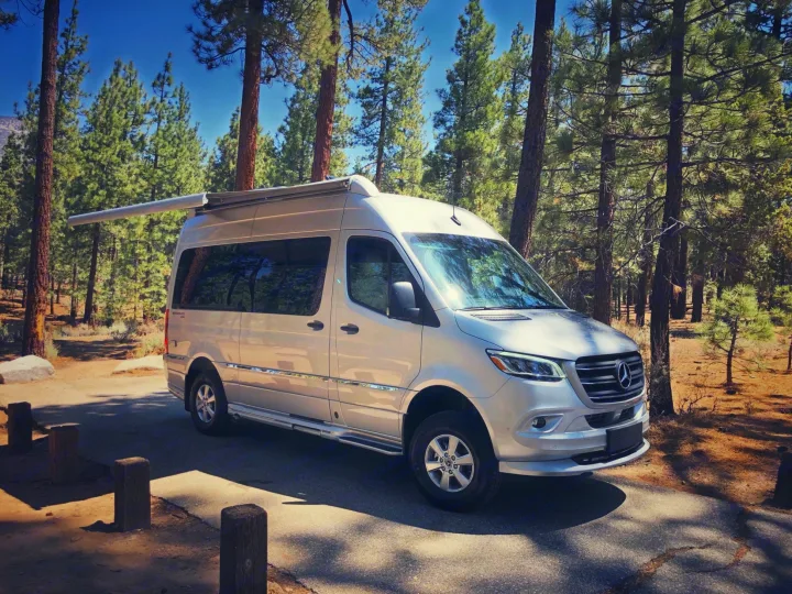 Silver Mercedes-Benz Sprinter camper van, an ideal RV rental, parked amidst tall pine trees in a sunny forest setting.. Airstream Interstate 2022