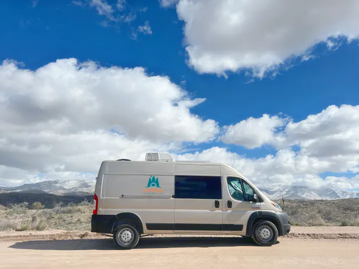 Scenic RV rental view: a tan camper van on a dirt road, under a blue sky with fluffy clouds & snow-capped mountains.. Dodge Promaster- Summit X Edition OA9 2021