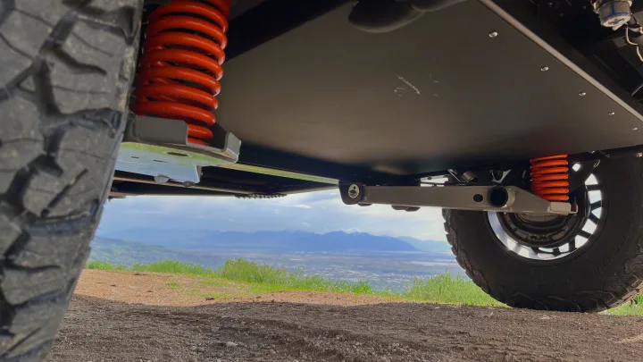 Underneath a rugged rv rental: orange springs, protective plates & chunky tires, seen from below, overlooking a mountain valley.. Other Other 2024