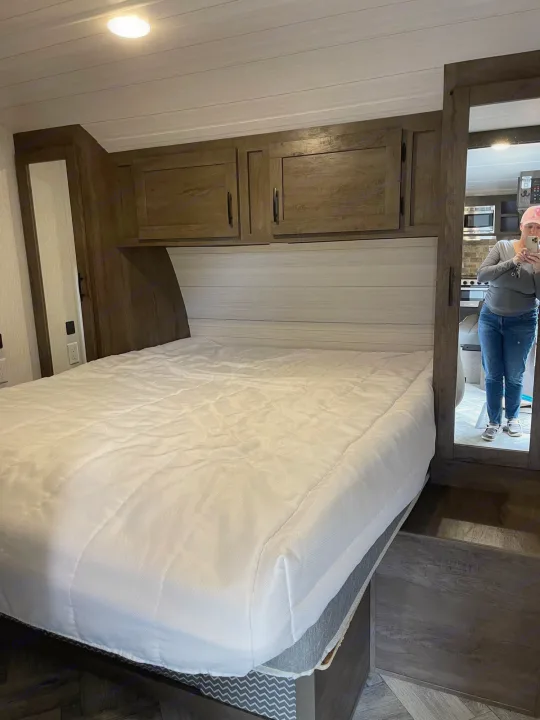 This RV rental bedroom features a cozy interior with a white-covered bed, overhead cabinets, and a mirrored view of the kitchen.. Forest River Evo 2022