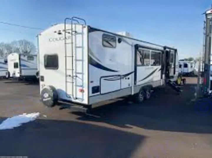 This white travel trailer, ready for RV rental, sits in a lot. Notice the rear ladder, extended slide-out, and awning.. Keystone Cougar 2023