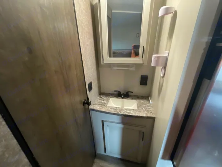 RV rental's compact bathroom with vanity, sink, faucet, mirror, wall storage, and a wooden door on the left.. Highland Ridge RV Open Range 2021