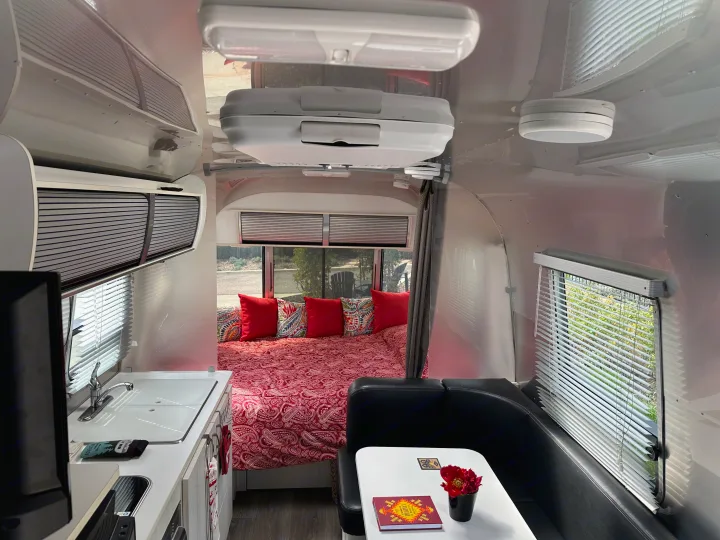 Inside a sleek RV rental: comfortable bed with red bedding, a dining nook, and a compact kitchen are visible.. Airstream Sport 2018