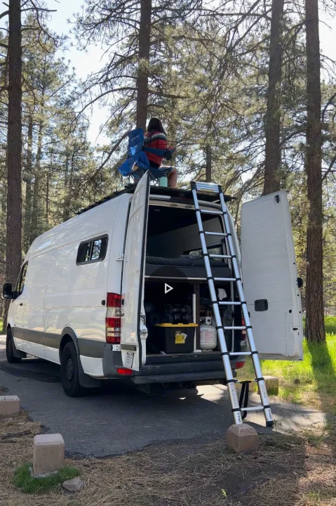 A person relaxes on the roof of a white RV rental camper van, parked in a sunny forest. A ladder extends from the open back doors.. Mercedes-Benz Sprinter 2013