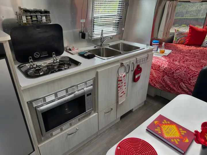 An RV rental kitchen, featuring a Dometic stovetop, Cuisinart microwave, and sink, next to a cozy bed.. Airstream Sport 2018