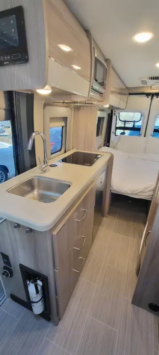 Modern RV rental interior: Compact kitchen with sink, cooktop, overhead storage, and a cozy bed area in back. Great for adventures!. Thor Motor Coach Sequence 2023