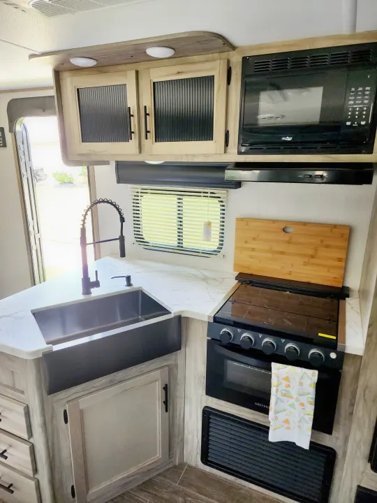 Bright RV rental kitchen: white marble-look counters, large sink, black faucet, microwave, oven/stove, and light wood cabinets.. Forest River Cherokee Alpha Wolf 2022