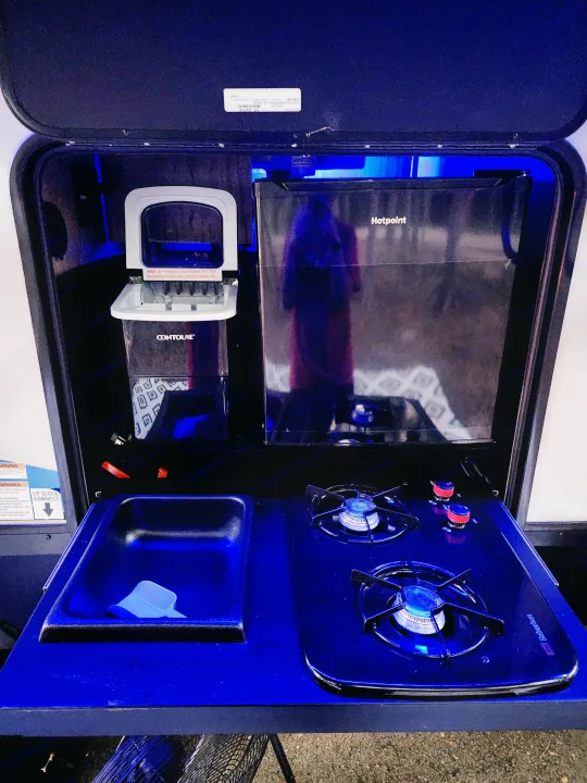 This **rv rental** outdoor kitchen features a Hotpoint fridge, Contoure ice maker, sink, and Suburban gas stove, all lit in blue.. Forest River Cherokee Alpha Wolf 2022
