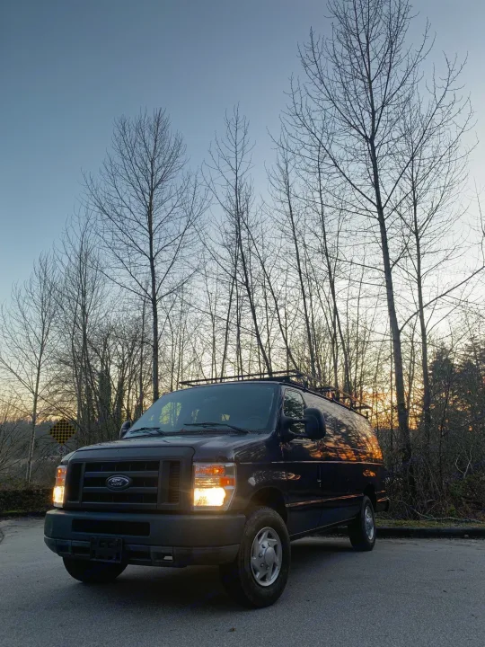 Ford van, possibly an rv rental, parked by bare trees silhouetted against a golden sunset. Headlights on.. Ford E350 Box Van 2010
