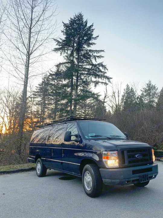 Dark camper van, an RV rental, parked roadside with tall trees silhouetted against the sunset.. Ford E350 Box Van 2010