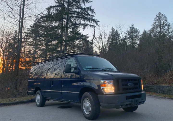 A dark Ford van, likely an rv rental, with a roof rack. Parked on asphalt near a forest at sunset with headlights on.. Ford E350 Box Van 2010