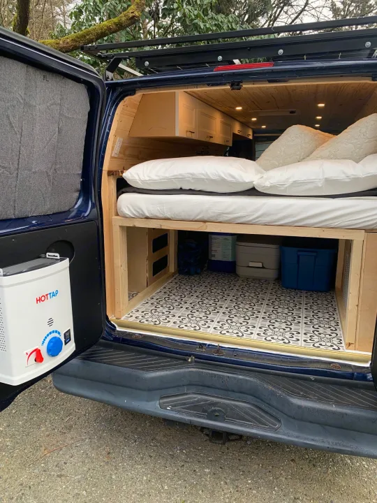 Rear view of an RV rental camper van interior: bed, tiled storage with toilet, & portable HOTTAP water heater setup.. Ford E350 Box Van 2010