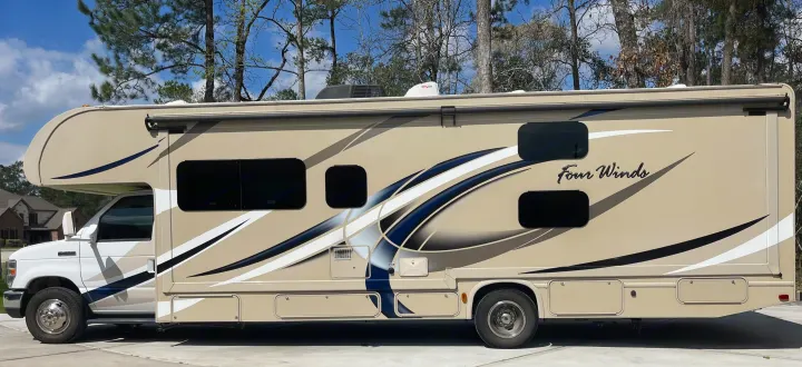 Side view of a tan Class C Four Winds RV rental, featuring blue and white graphics, parked outdoors. Ready for travel.. Thor Four Winds 2017