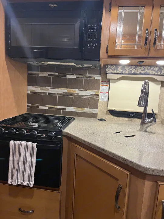 This RV rental kitchen features a black Whirlpool microwave, Wedgewood Vision stove, sink, and tiled backsplash for convenient travel meals.. Thor Four Winds 2017