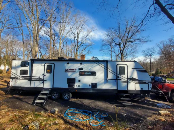 This white Coleman travel trailer (RV) is parked on a driveway, featuring two doors & steps, ready for RV rental adventures!. Dutchmen Coleman Light 2021