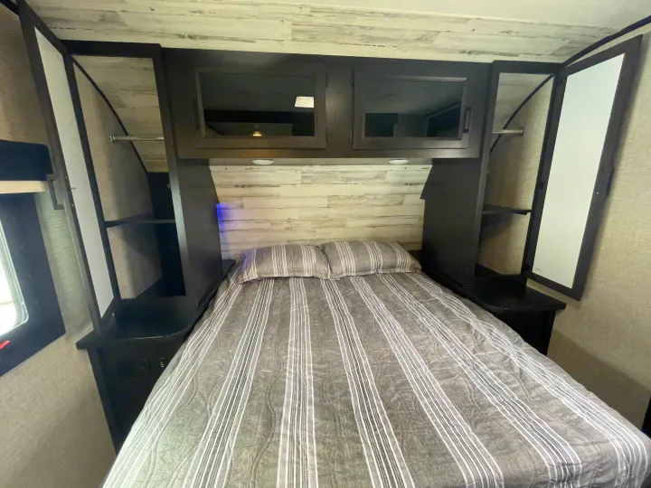 Comfortable RV rental bedroom featuring striped bedding, overhead cabinets, and convenient side storage.. Jayco Jay Feather 2022