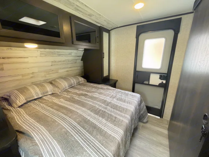 Cozy RV rental bedroom featuring a bed with striped linens, wood-paneled accent wall, overhead storage, and a side door.. Jayco Jay Feather 2022