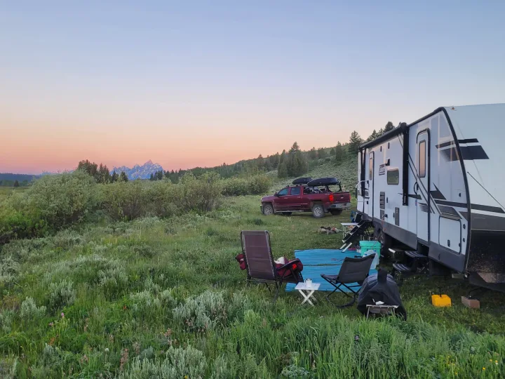 Beautiful RV rental camping scene: travel trailer, truck, and chairs in a grassy field, mountains at sunset.. Dutchmen Coleman Light 2021