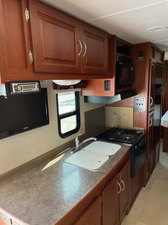 Inside an rv rental: a compact, functional kitchen with dark wood cabinets, sink, stove, microwave, and integrated entertainment.. Winnebago Minnie Winnie 2015