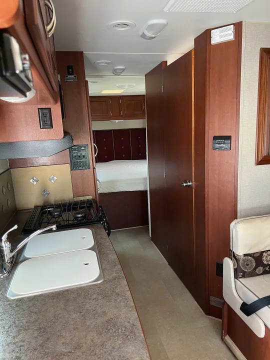 This RV rental interior features a compact kitchen with a sink and stove, a peek into the bedroom, and accessible controls.. Winnebago Minnie Winnie 2015