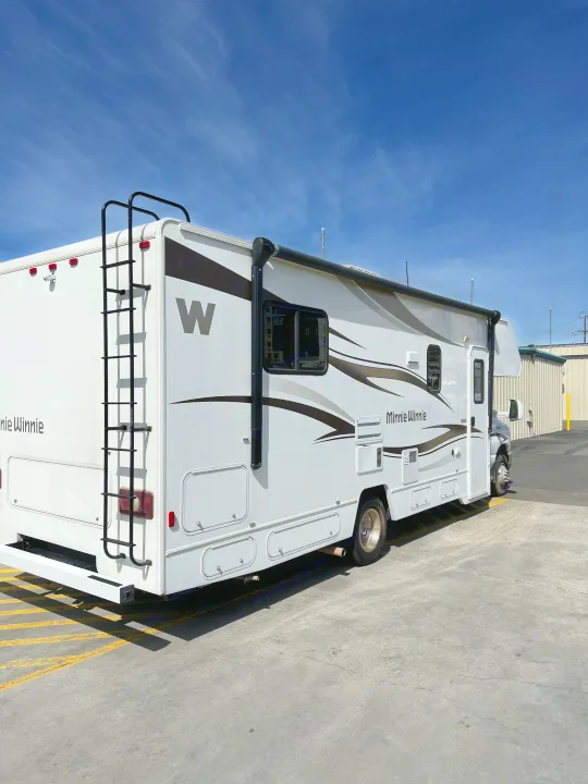 White Minnie Winnie RV rental parked outdoors, showing side details, rear ladder, and blue sky.. Winnebago Minnie Winnie 2015