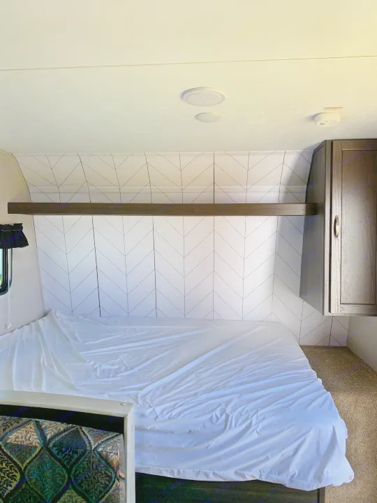 Inside an RV rental bedroom: white bed, black & white chevron headboard, wood shelf & cabinet, white ceiling with speakers.. Keystone Springdale 1800BH 2019