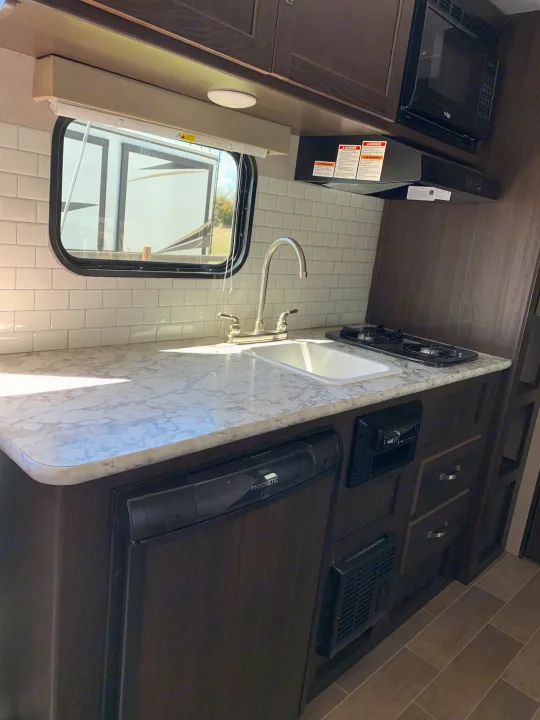 Compact kitchen in RV rental, showing sink, stove, microwave, and fridge with modern tile backsplash.. Keystone Springdale 1800BH 2019