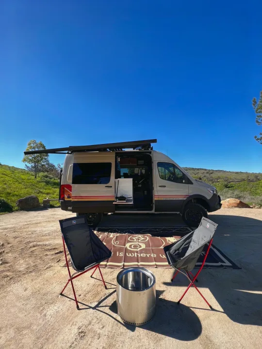 Perfect rv rental setup: camper van, chairs, Solo Stove fire pit on dirt with clear blue sky & green hills for an adventure.. Storyteller Overland Beast Mode 2023