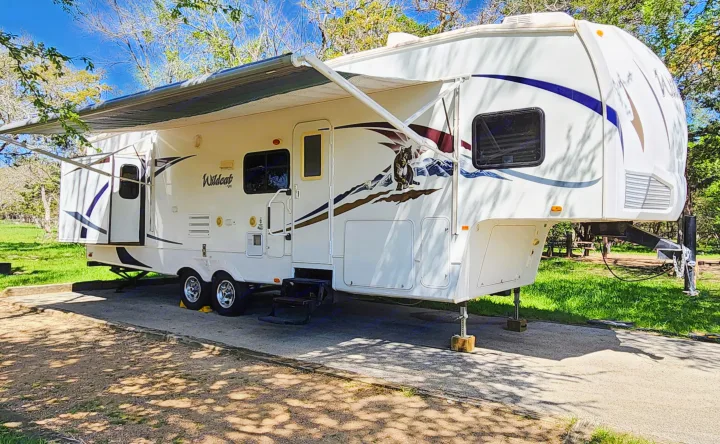 White fifth-wheel RV rental with extended awning, parked on a concrete pad in a sunny, tree-lined campsite.. Forest River Wildcat 2008