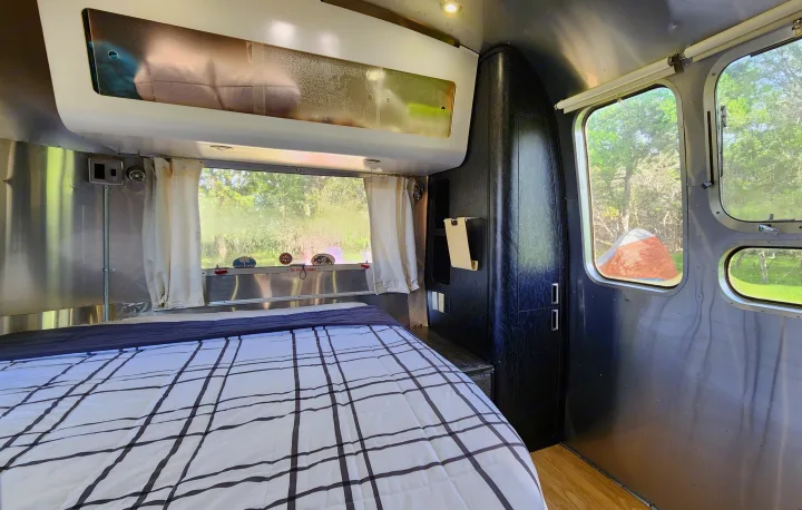 Interior of a shiny Airstream RV rental bedroom: large bed with plaid comforter, big windows showing green trees.. Airstream International 2004