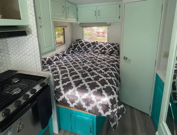 Compact RV rental interior featuring a bed with patterned comforter, light green cabinets, and a kitchen stove area.. Other Other 2005
