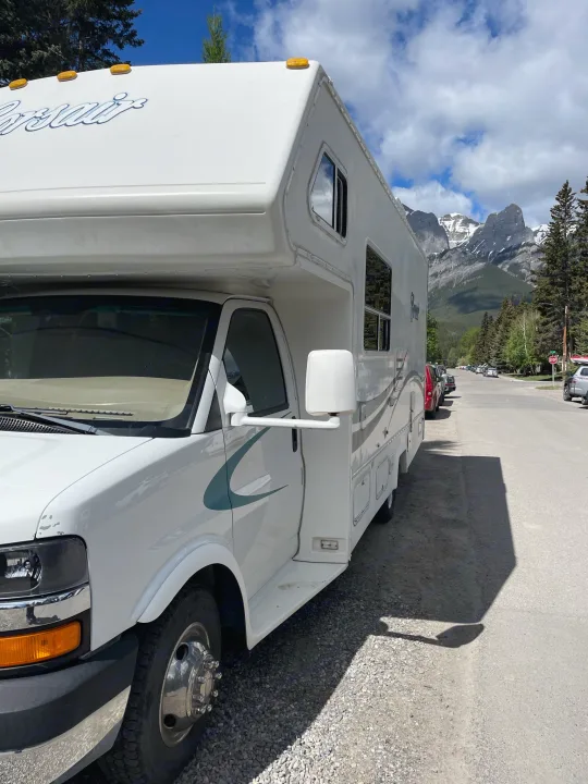 This white RV rental is parked by a road, framed by majestic snow-capped mountains and green trees under a blue sky.. Other Other 2005