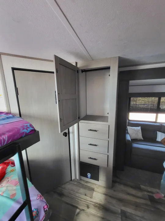 RV rental interior: light wood cabinets, sofa, window with blinds, and bunk area with colorful bedding. Practical and bright space.. Dutchmen 285BH 2022