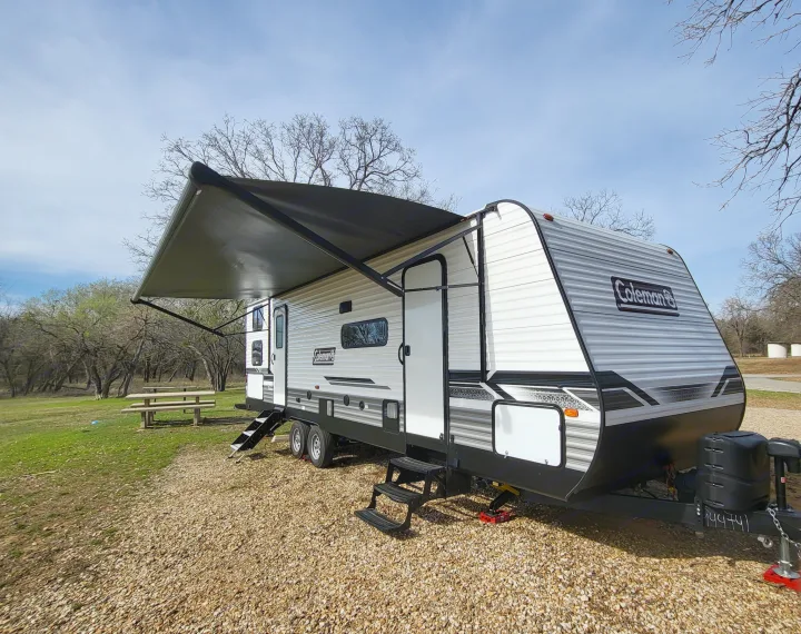 A sleek Coleman travel trailer, ready for RV rental, with its awning extended at a gravel campsite surrounded by trees.. Dutchmen 285BH 2022