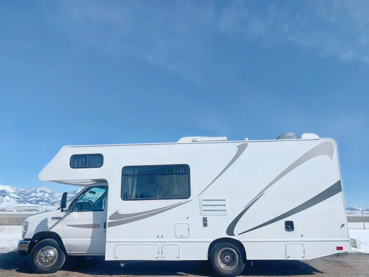 White Class C RV rental with gray stripes parked against a clear blue sky, snow-capped mountains.. Thor Freedom Elite 2019