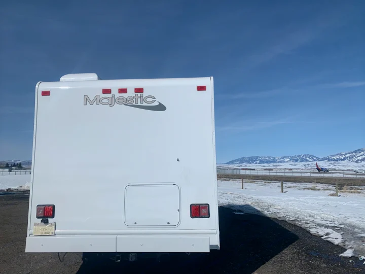 Rear view of a white Majestic RV rental parked in a snowy landscape, with mountains and a Southwest plane at an airport in the distance.. Thor Freedom Elite 2019