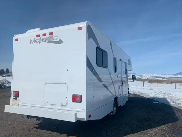 Majestic RV rental, white with gray accents, parked on a gravel lot. Snowy mountains and a blue sky in the background.. Thor Freedom Elite 2019