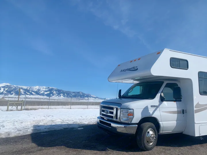 White Majestic Ford E-350 rv rental parked on a partly snowy lot, facing snow-capped mountains under a clear blue sky.. Thor Freedom Elite 2019