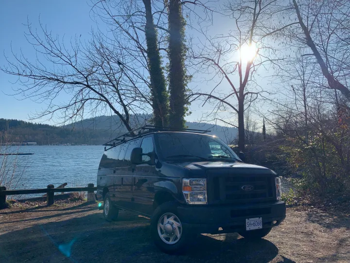 Black Ford E-Series van, perfect for RV rental, parked by a lake with mountains under a bright sky.. Ford E350 Box Van 2010