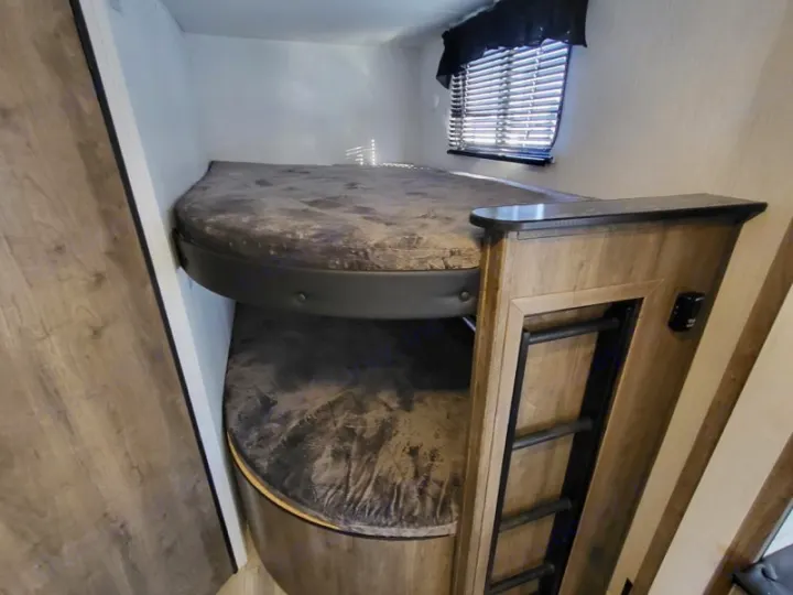 Inside an RV rental: two compact bunk beds with rounded ends, a ladder for the top, and a window with blinds.. Heartland Pioneer BH270 2023