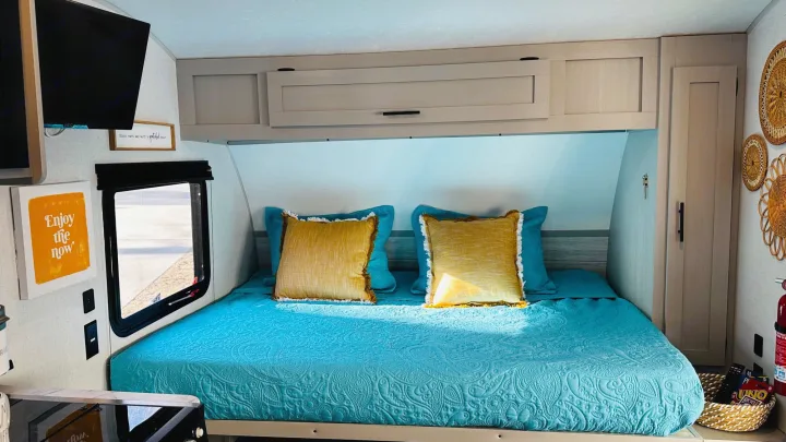Inside a cozy RV rental: a bright blue bed with yellow pillows, overhead cabinets, TV, window, and woven wall decor.. Forest River Wildwood 2021