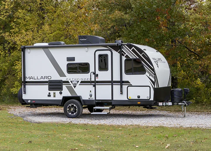 A white Mallard Pathfinder travel trailer, suitable for an RV rental, parked on gravel in a wooded area.. Heartland RVs Mallard 2022