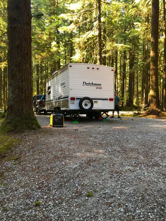Scenic RV rental campsite in a tall forest, featuring a white travel trailer, truck, and generator.. Dutchmen Sport 2004